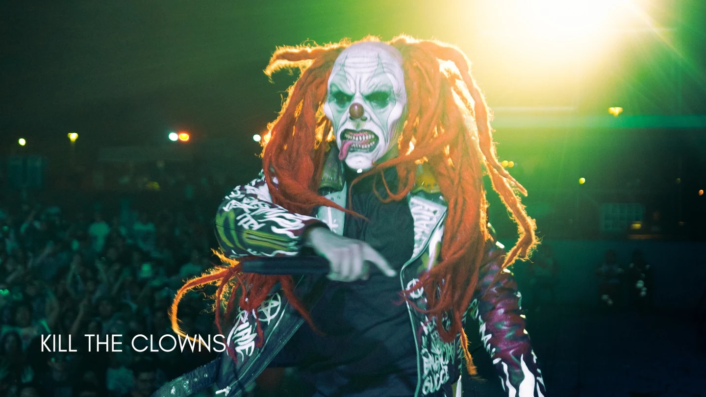 Artista de KILL THE CLOWNS con máscara y cabello naranja durante un concierto, entre luces verdes y amarillas.